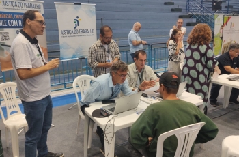 Foto de um atendimento feito por um homem a outro homem, em mesas de plásticos, dentro de um ginásio