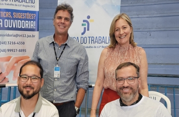 Quatro pessoas sorriem para a foto. Dois homens, sentados; e duas pessoas em pé, sendo um homem e uma mulher. Ao fundo, um banner vertical onde se lê "Justiça do Trabalho"