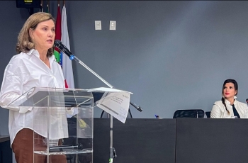 Mulher fala ao microfone em púlpito; outra mulher sentada à mesa a observa, em ambiente formal com bandeiras ao fundo.