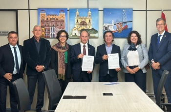 Duas mulheres e cinco homens estão em pé, sorrindo e posando para a foto, em uma sala de reuniões com quadros coloridos na parede