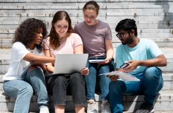 Grupo de quatro estudantes sentados em escadaria ao ar livre, olhando para um notebook com atenção.