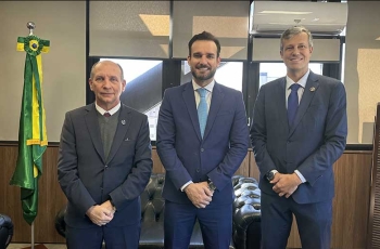 Três homens de terno, lado a lado, posam para foto, dentro de uma sala. À esquerda, aparece a bandeira do Brasil