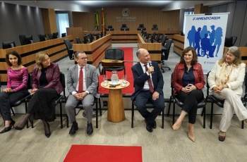 Quatro mulheres e dois homens sentados à frente de um auditório, em cadeiras. Ao fundo, um violão e um banner escrito Aprendizagem