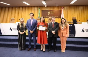 Fotografia na qual cinco pessoas posam em auditório do TST; ao centro, mulher segura certificado