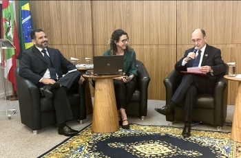Dois homens de terno e uma mulher de camisa verde estão sentados dando uma palestra em um evento