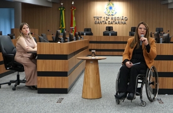 Foto de uma mulher de cadeiras de rodas dando uma palestra. No fundo um painel escrito Tribunal Regional do Trabalho da 12ª Região.