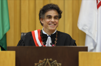 Homem de cerca de 60 anos, vestido com toga de juiz e faixa, sorri 