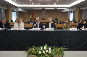 Três homens e três mulheres sentadas à mesa de um evento, coberta por uma toalha preta e um buquê de flores no chão.