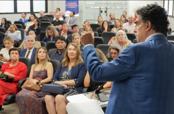 Foto de um homem de costas, de terno, dando uma palestra. Ao fundo o público assistindo