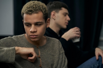 Fotografia ilustrativa de homem jovem pensativo em primeiro plano, outro desfocado ao fundo segurando copo