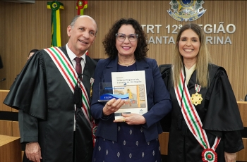 Três pessoas sorridentes posam em solenidade no TRT-12; ao centro, mulher segura troféu e livro comemorativo.