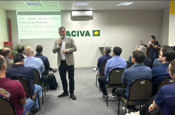 Um homem esta em pé fazendo uma palestra em uma sala de eventos, onde homens e mulheres estão sentados assistindo