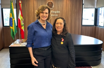 Duas mulheres, uma loira e outra morena, posam sorridentes para a foto. A morena tem uma medalha fixada na roupa