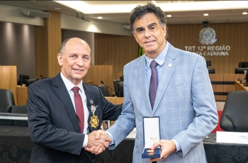 Dois homens de terno, com cerca de 60 anos, se cuprimentam. Um deles, de terno claro, exibe uma caixa contendo uma medalha.