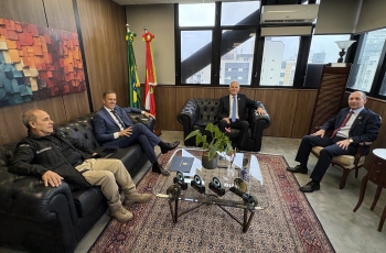 Três homens de terno e um com uniforme de segurança sentados em poltronas e cadeiras em uma sala arejada