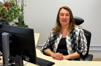 Mulher de pele branca e cabelos castanhos claros, sentada em um posto de trabalho, sorri para a câmera