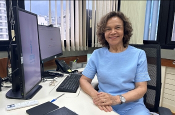 Mulher parda, madura, de conjunto azul claro e cabelos castanhos sentada à mesa de trabalho