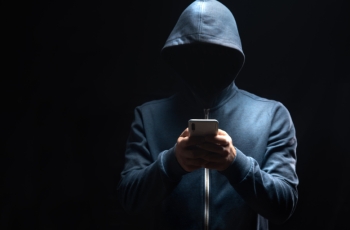 Pessoa de moletom com capuz, rosto oculto, usando celular em ambiente escuro