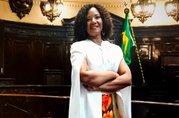 Mulher negra, de pé, de braços cruzados, em uma sala de julgamentos