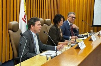 Foto registrada em ângulo de 45 graus mostra dois homens de terno e uma mulher sentados à mesa de um evento