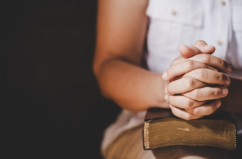 Fotografia ilustrativa de mulher com mãos entrelaçadas, apoiadas sobre um livro fechado