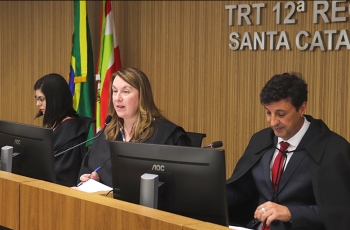 Duas mulheres e um homem sentados atrás de uma bancada. Ao fundo, bandeiras do Brasil e de SC e um letreiro escrito TRT 12ª Região - Santa Catarina