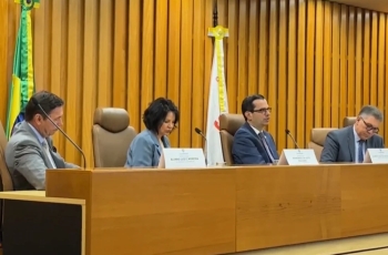 Três homens de terno e uma mulher vestida formalmente sentados em uma mesa de eventos, em tom madeira-ouro, com uma bandeira do Brasil atrás