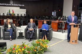 Duas mulheres e um homem sentados em poltronas e um quarto homem, ao lado, falando em um púlpito no auditório do Tribunal de Justiça de SCo