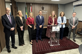 Três homens e quatro mulheres em pé, em uma sala, lado a lado, todos sorridentes. Uma das mulheres assina um documento em um púlpito transparente