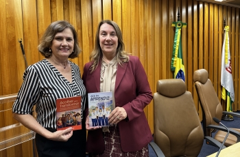 Duas mulheres posam para a foto. Cada uma exibe uma cartilha