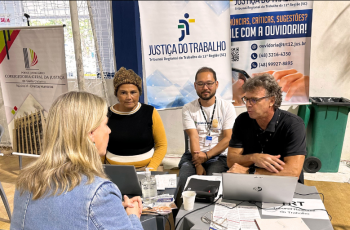 Foto de dois homens e uma mulher prestando atendimento a uma mulher loira. Atrás da mesa, um banner da Justiça do Trabalho