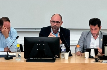 Homem calvo, de óculos, sentado à frente de um notebook, faz uma apresentação. Ao lado dele estão outros dois homens.