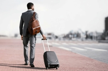 Fotografia ilustrativa de homem negro de terno claro e malas olhando para o horizonte distante