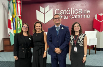 Três mulheres vestidas formalmente e um homem de terno azul marinho posam para a foto, sorridentes, em um auditório. Ao fundo, a logomarca da Universidade Católica