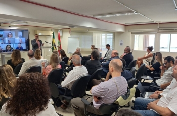 Homem dá palestra a público sentado em sala; bandeiras ao fundo e telão com videoconferência