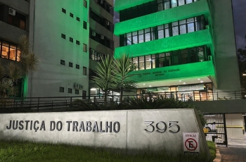 Parte da fachada do TRT-SC iluminada de verde, mostrando três pavimentos. À frente, no chão, um letreiro escrito Justiça do Trabalho