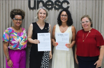 Quatro mulheres estão em pé, posando e sorrindo para a foto, segurando um contrato