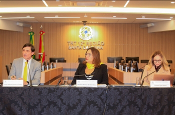 Um homem e duas mulheres estão sentados em uma mesa de honra, durante uma palestra