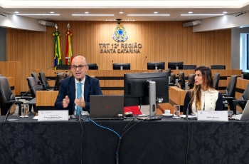Duas pessoas, um homem e uma mulher, sentados diante de mesa de honra durante palestra em plenário