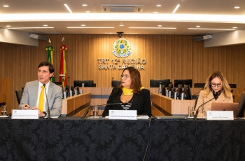 Um homem e duas mulheres estão sentados em uma mesa de honra, durante uma palestra