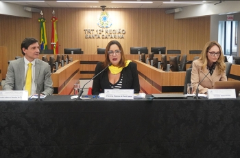 Um homem e duas mulheres estão sentados em uma mesa de honra, durante uma palestra