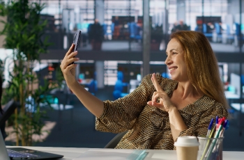 Fotografia ilustrativa de mulher sorrindo para o próprio celular em ambiente de escritório