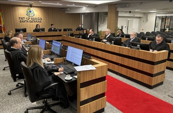 13 pessoas vestem roupas pretas, todas sentadas em torno de duas bancadas laterais e uma central, formando a letra U