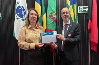 Uma mulher vestida formalmente e um homem de terno e gravata seguram, juntos, um certificado. Ao fundo bandeiras do Brasil e dos estados do Paraná, Santa Catarina e outra não identificada
