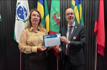 Uma mulher vestida formalmente e um homem de terno e gravata seguram, juntos, um certificado. Ao fundo bandeiras do Brasil e dos estados do Paraná, Santa Catarina e outra não identificada