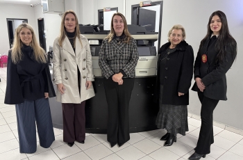 Cinco mulheres posam para a foto. Atrás delas, dois equipamentos de informática