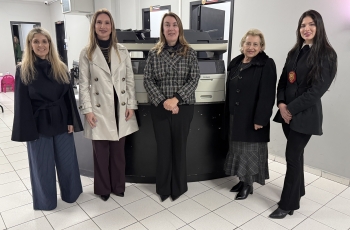Cinco mulheres posam para a foto. Atrás delas, dois equipamentos de informática