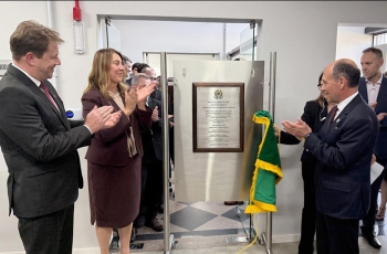 Dois homens de terno e gravata e uma mulher estão aplaudindo felizes em frente a uma placa de inaguração de um forum trabalhista