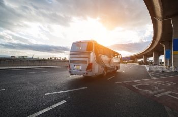 onibus branco, em uma estrada, com o céu de fundo possuindo sol e nuvens
