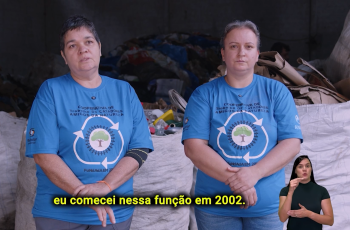 Duas mulheres brancas, uma de cabelos escuros e outra loira, estão lado a lado, olhando para a câmera, à frente de resíduos para reciclagem
